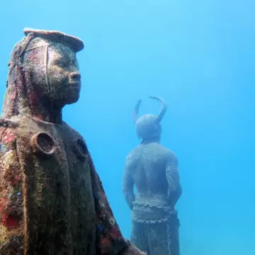 Two submerged statues coated in coral growth standing in bright blue water, one robed and facing the camera, one horned and turned away.