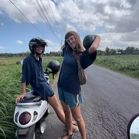 Two women holding watermelons stand next to a motorbike. 