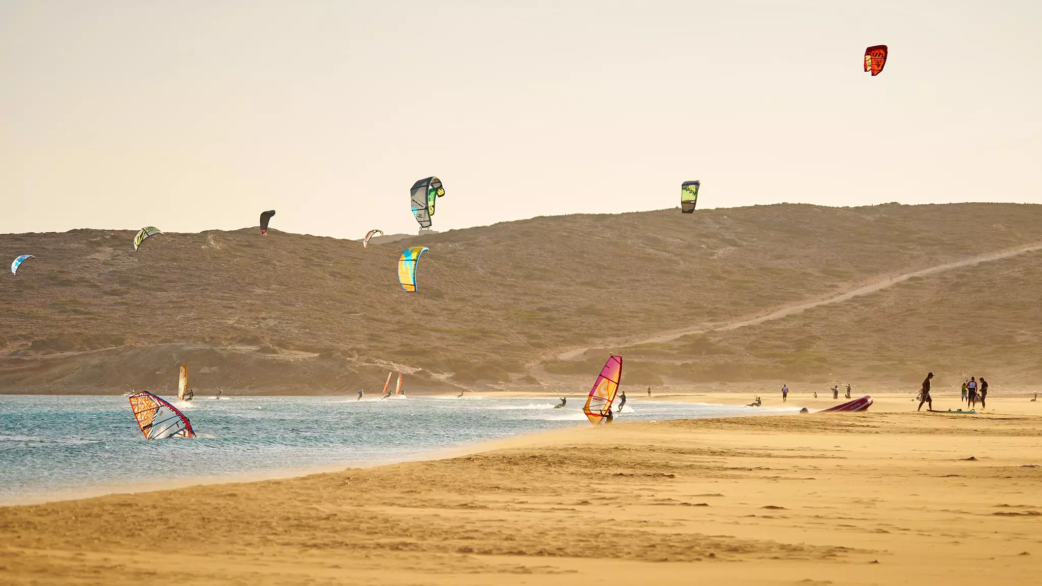 Windsurfers and kitesurfers are pictured in hazy light at a beach.