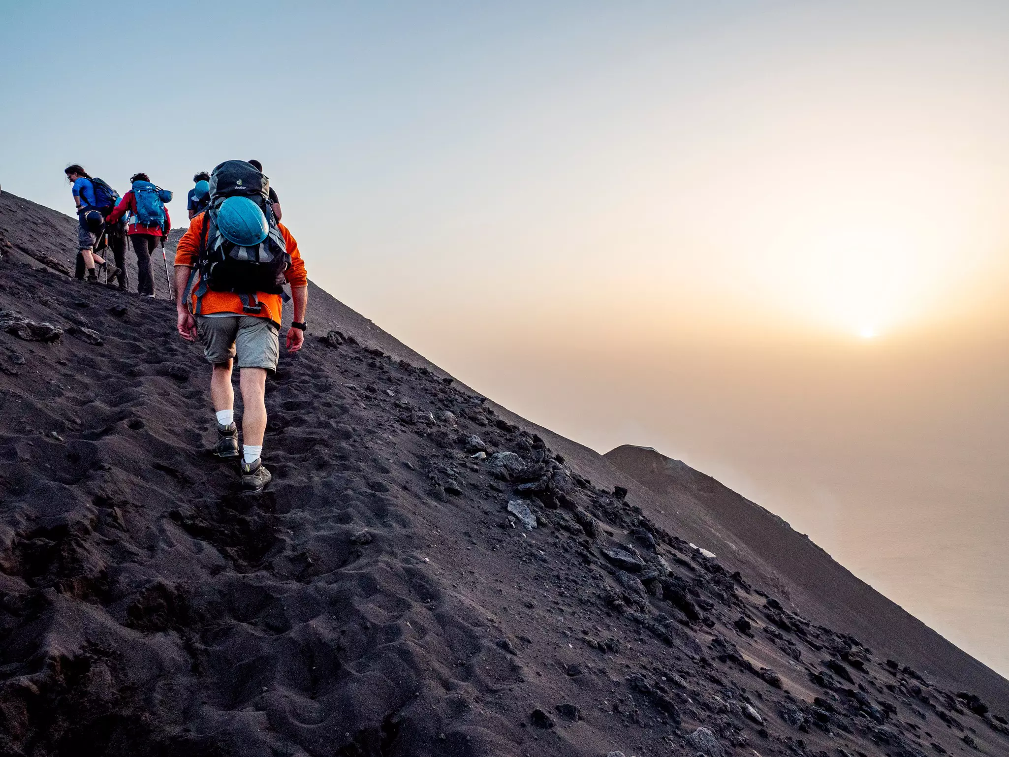 Hikers with backpacks and trekking poles walking along a volcano's ash-covered path at sunset.