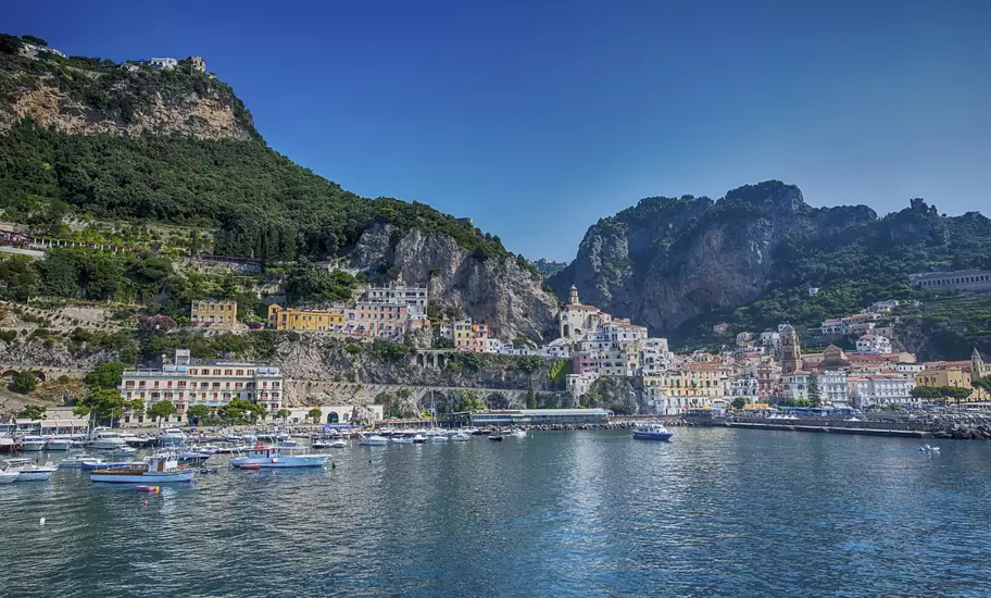 Amalfi town rising from its harbour, moored boats and pastel buildings below green cliffs and the cathedral bell tower