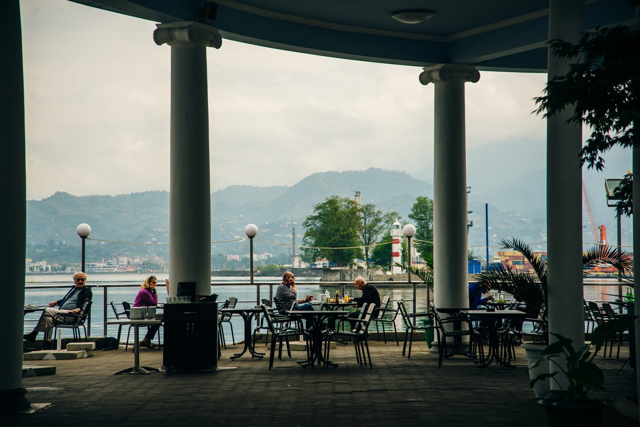 Diners at a colonnaded waterfront cafe in Batumi facing the red-and-white lighthouse, port cranes and green hills