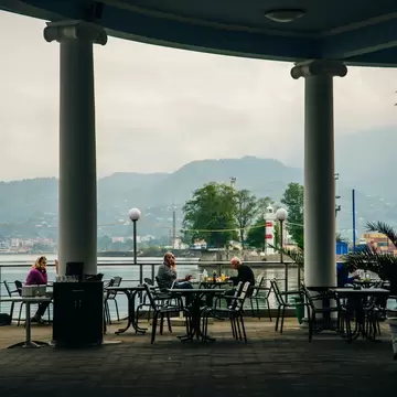 Diners at a colonnaded waterfront cafe in Batumi facing the red-and-white lighthouse, port cranes and green hills