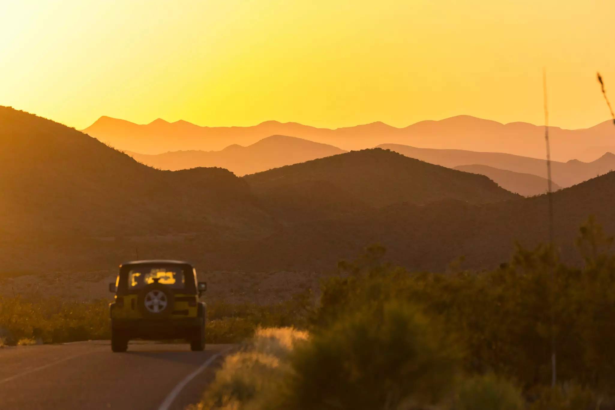 A car is seen from behind driving down a road during sunset. Golden light illuminates ridges of mountains in the distance.