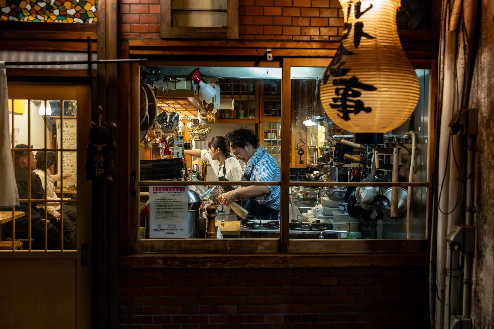Chefs at a restaurant are seen preparing a meal through a window from the street in a city at night