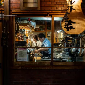 Chefs at a restaurant are seen preparing a meal through a window from the street in a city at night