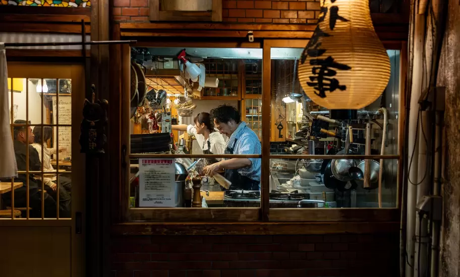 Chefs at a restaurant are seen preparing a meal through a window from the street in a city at night