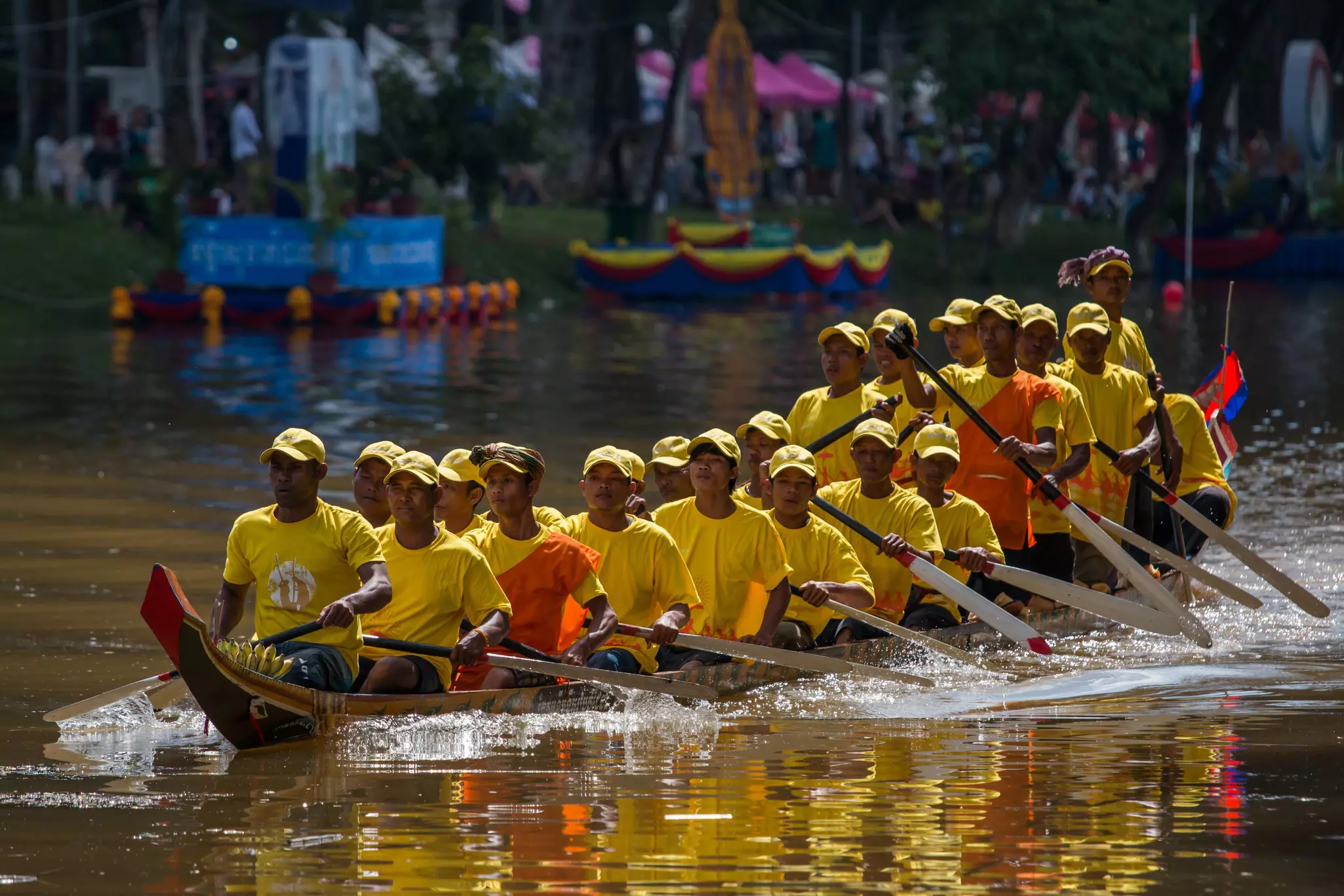 Rowers in a traditional boat wear yellow T-shirts and hats while paddling on a river in Siem Reap, Cambodia.