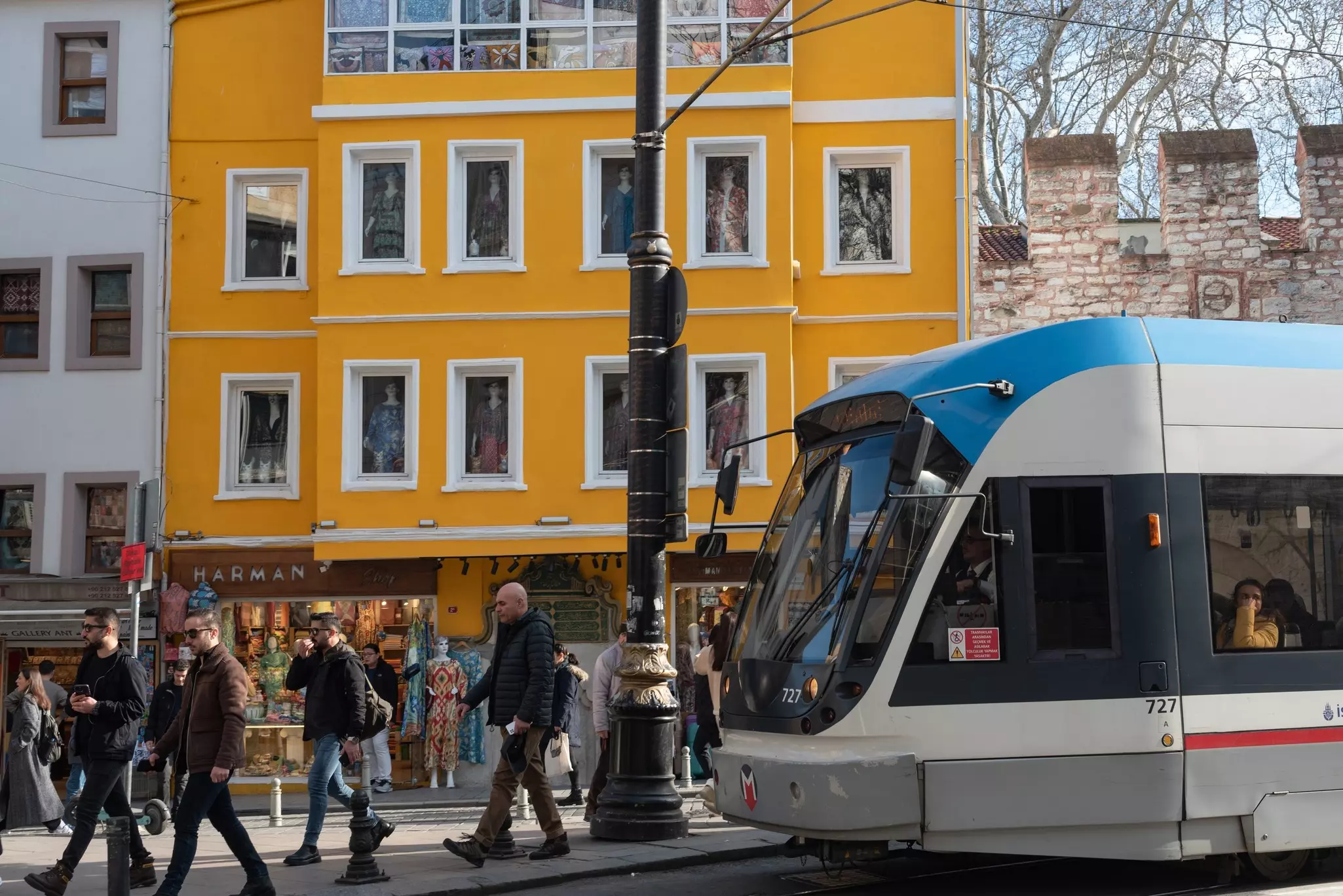 People walking on a sidewalk near a blue, gray and white tram that is stopped in front of a bright yellow building