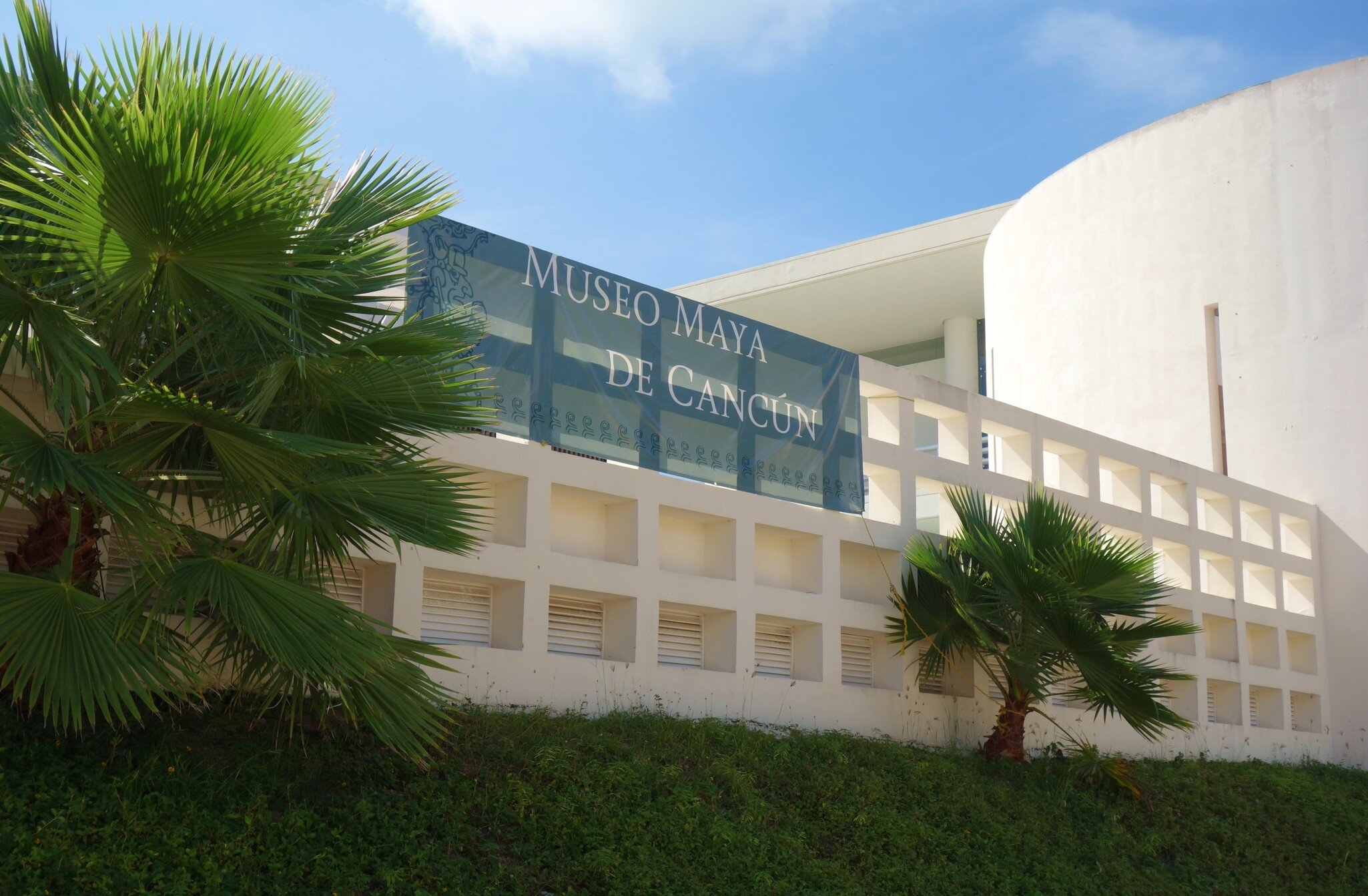 Blue banner lettered "Museo Maya de Cancún" on a white lattice wall, palm fronds across the frame under a clear sky.