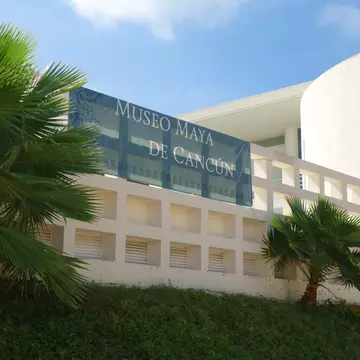 Blue banner lettered "Museo Maya de Cancún" on a white lattice wall, palm fronds across the frame under a clear sky.
