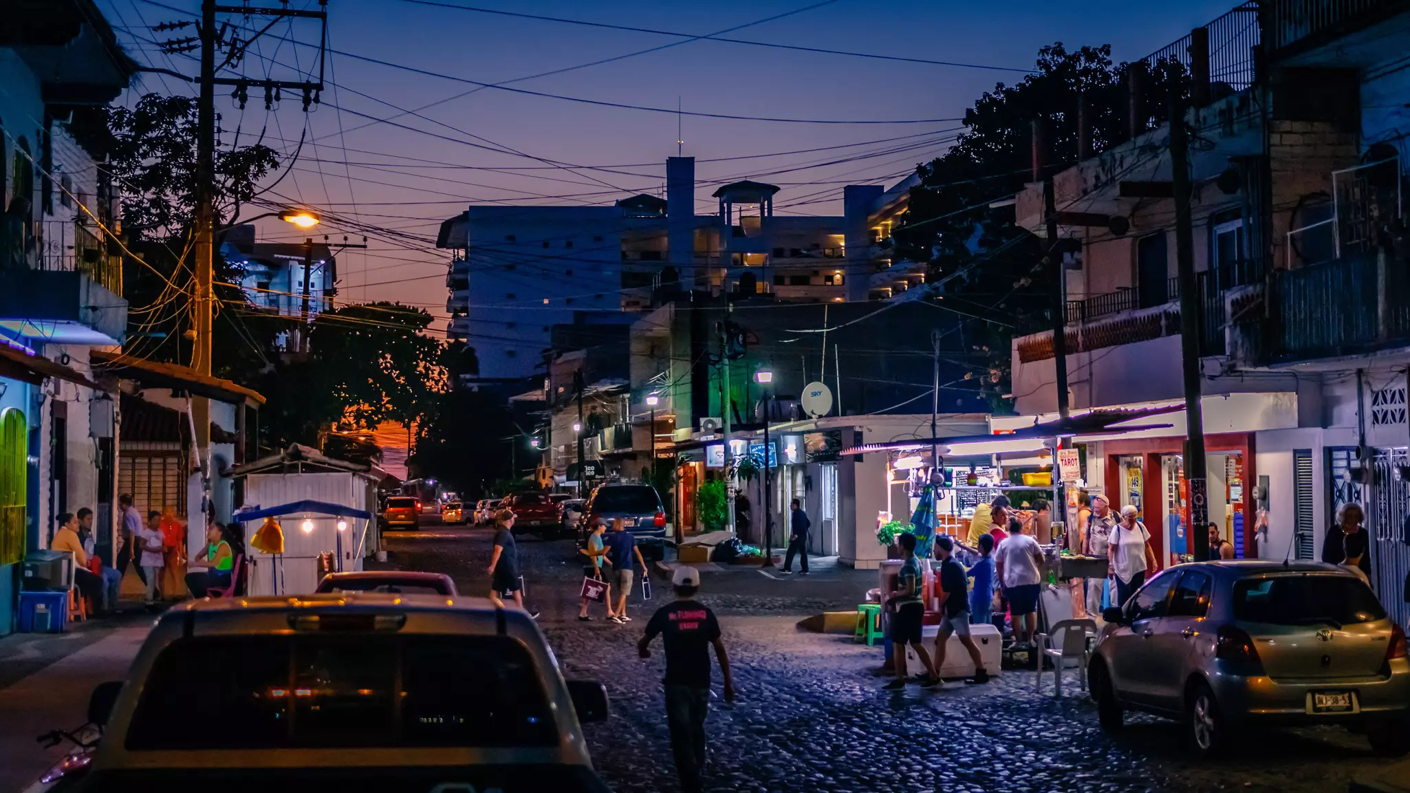 People walk at sunset on a cobblestone street in a city, with the lights of restaurants and businesses illuminating the scene