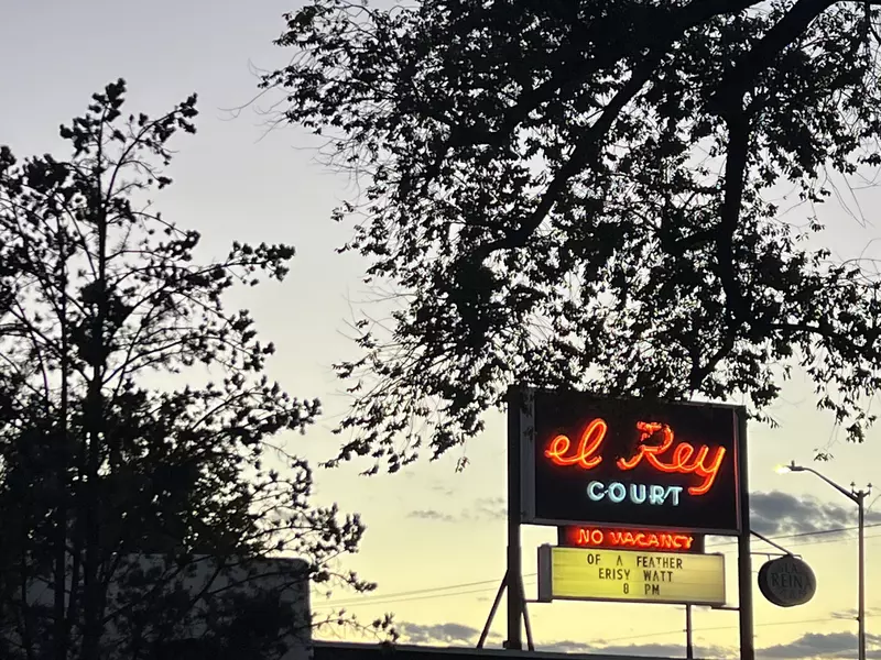 A neon sign reading "El Rey Court" set against the silhouette of tree leaves at sunset.