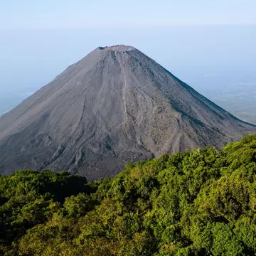 The gray dusty surface of the cone of a volcano surrounded by dense jungle.