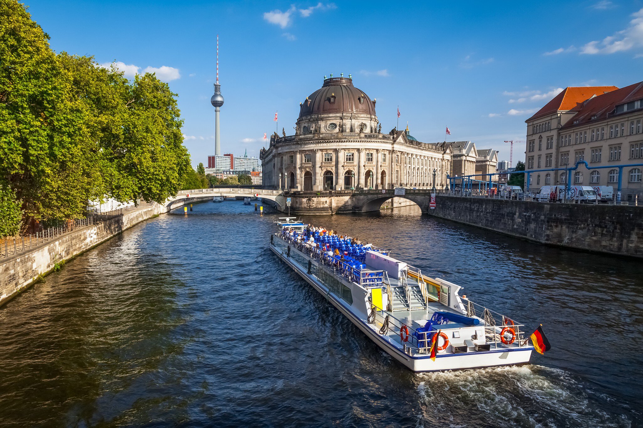A white tour boat with rows of blue seats and a German flag cruises the Spree past the domed Bode Museum.