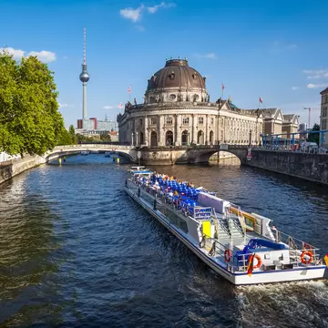 A white tour boat with rows of blue seats and a German flag cruises the Spree past the domed Bode Museum.
