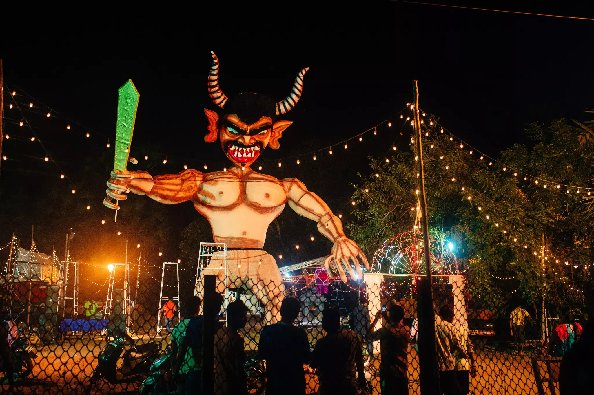 A handmade effigy of a demon rises over a crowd at night, surrounded by strings of lights.