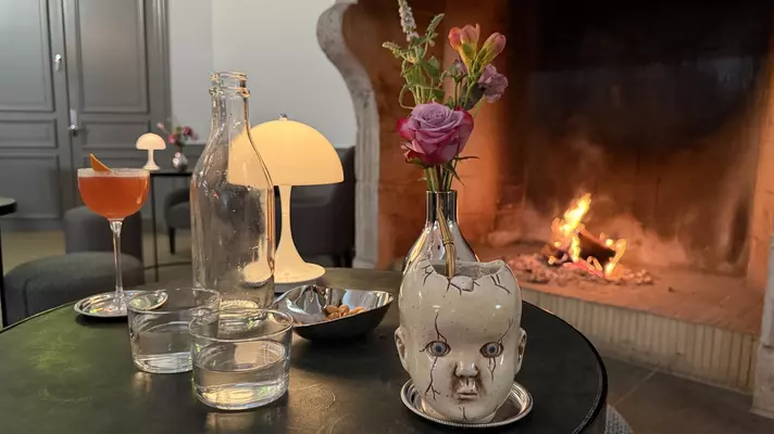 Close-up of a cocktail mug that looks like a baby's head on a table alongside other glasses and a bottle, with fireplace in the background.