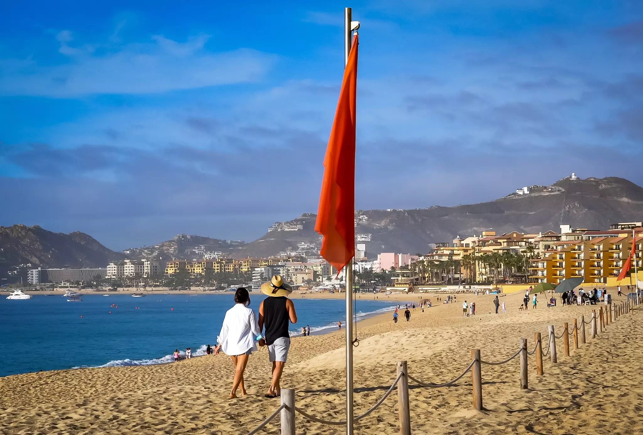 Two people walk past a closed red umbrella on a sandy beach with development at the far end.