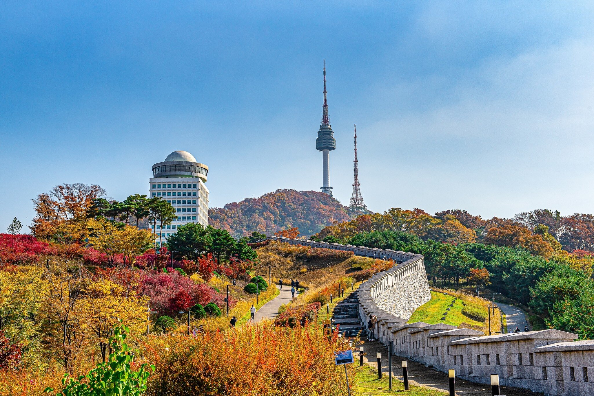 Stone fortress wall and walking paths lined with red and yellow foliage lead up a hill topped by a tall tower