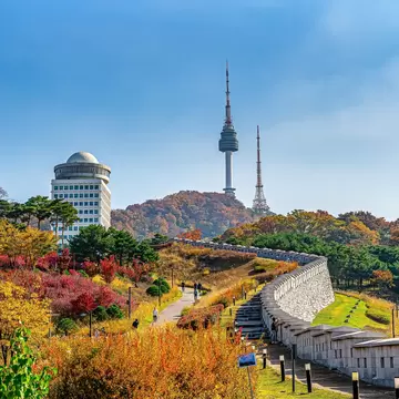 Stone fortress wall and walking paths lined with red and yellow foliage lead up a hill topped by a tall tower