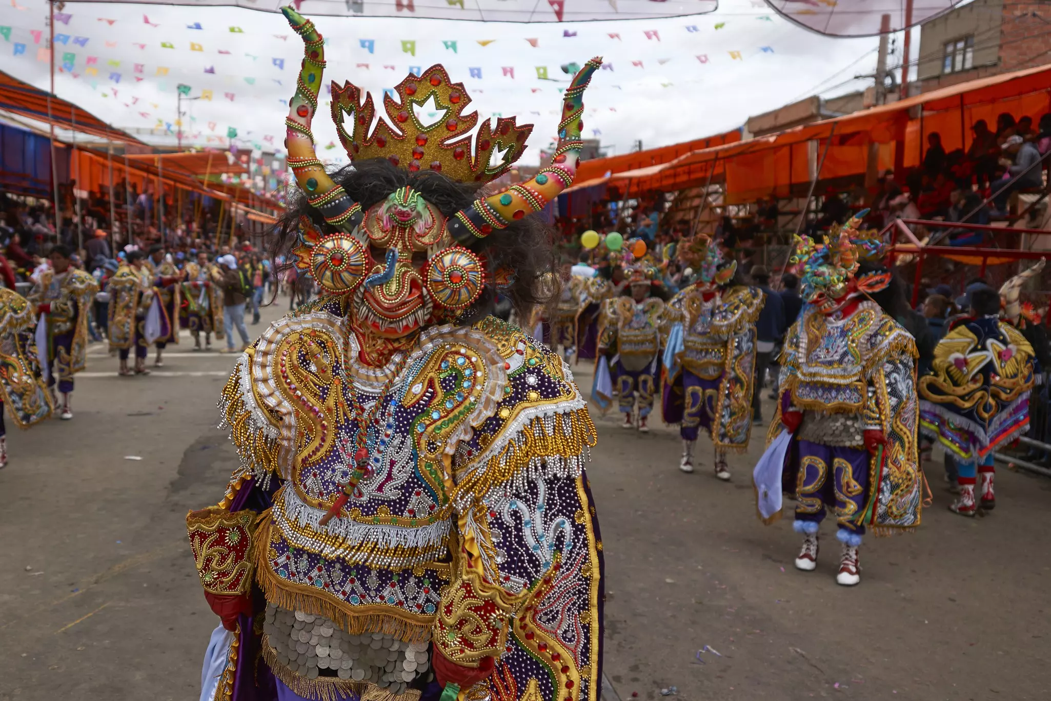Dancers in ornate costumes covered in beads and tassles wear elaborate masks with horns.