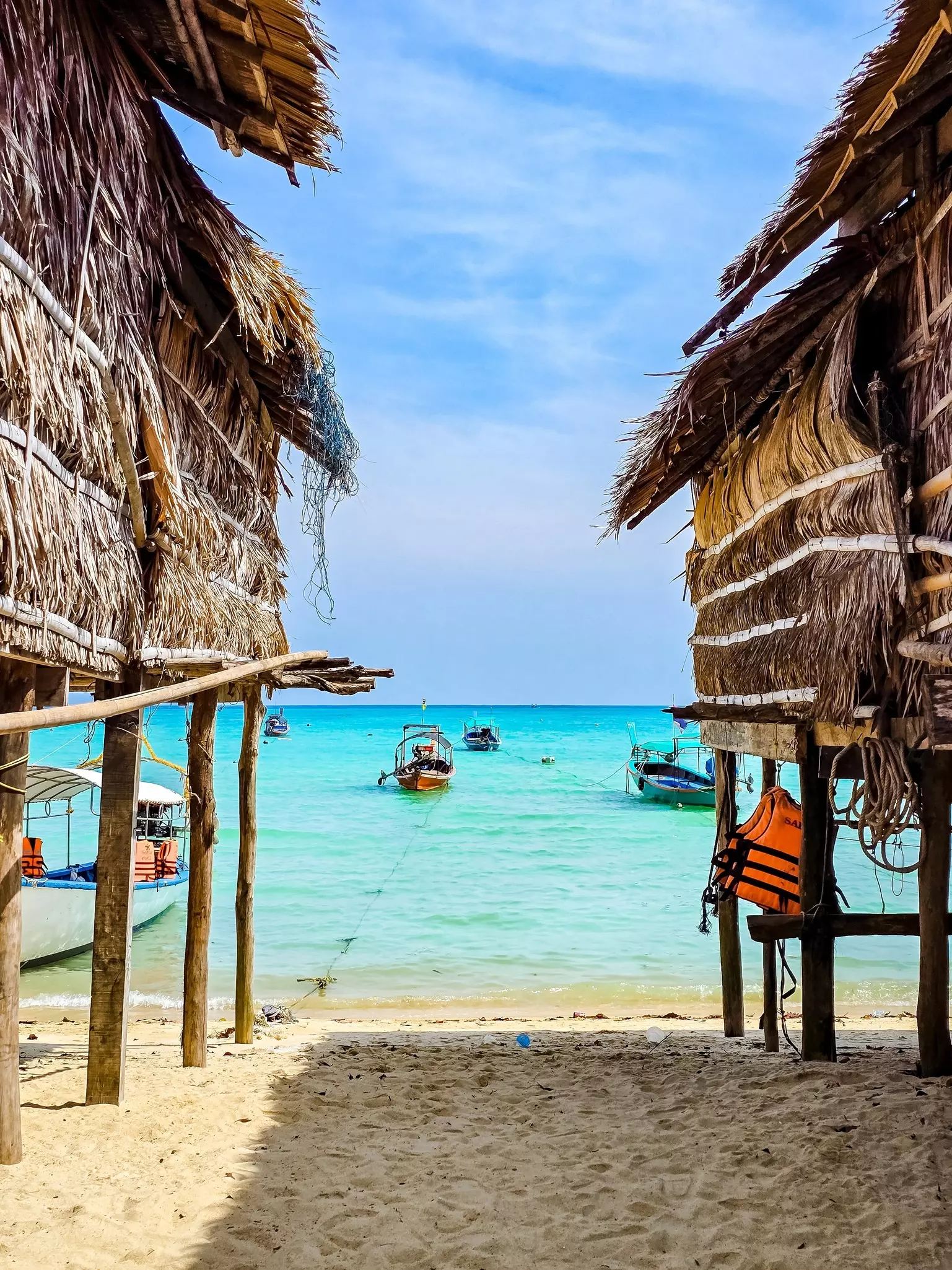 Crystal-clear turquoise waters embrace the sandy shore of an island, where colorful boats rest peacefully. Rustic straw huts frame the breathtaking view, inviting tranquility and exploration