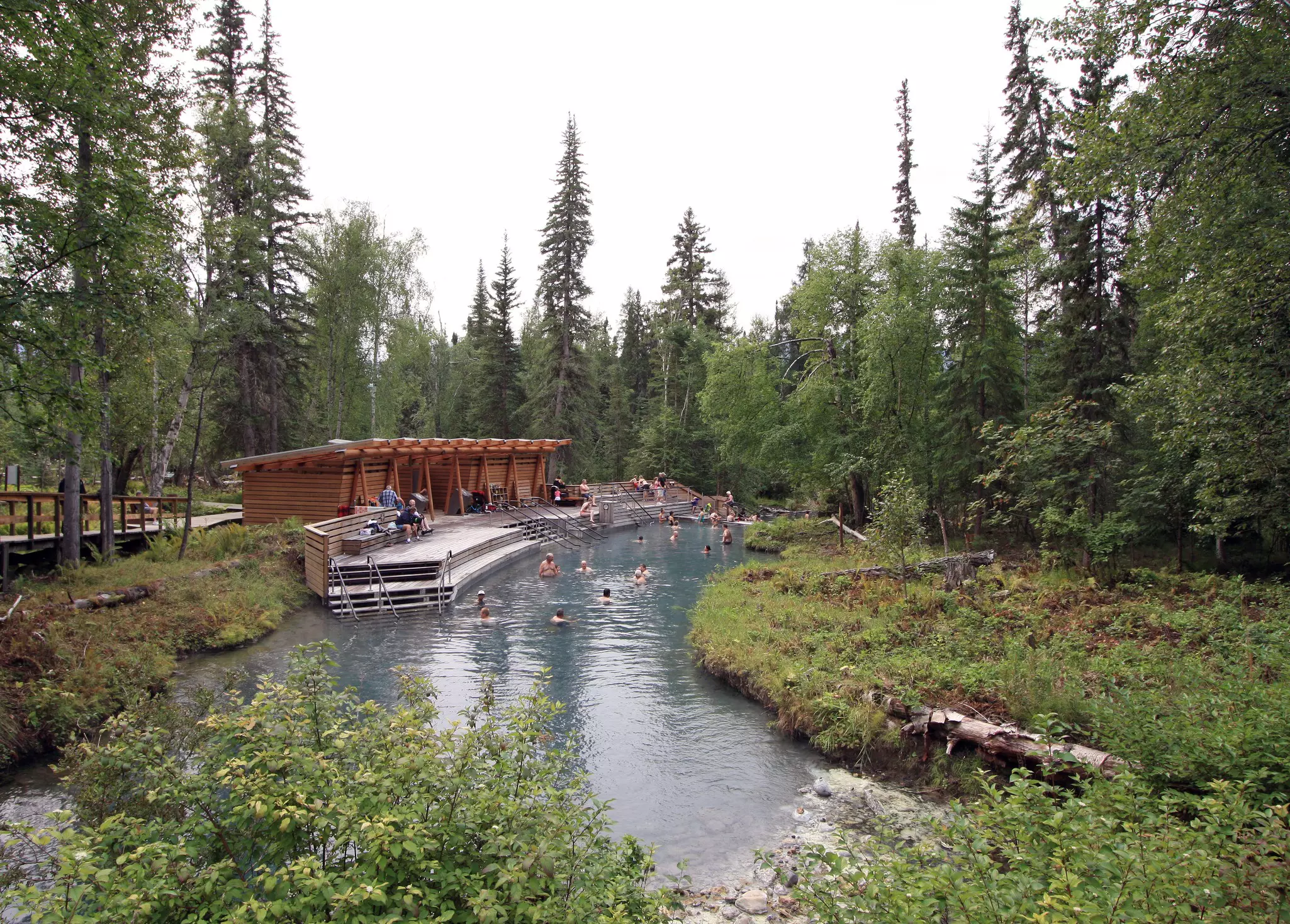 People soak in a river hot spring with stairs leading to a wooden patio and covered bathhouses surrounded by evergreens on an overcast day.