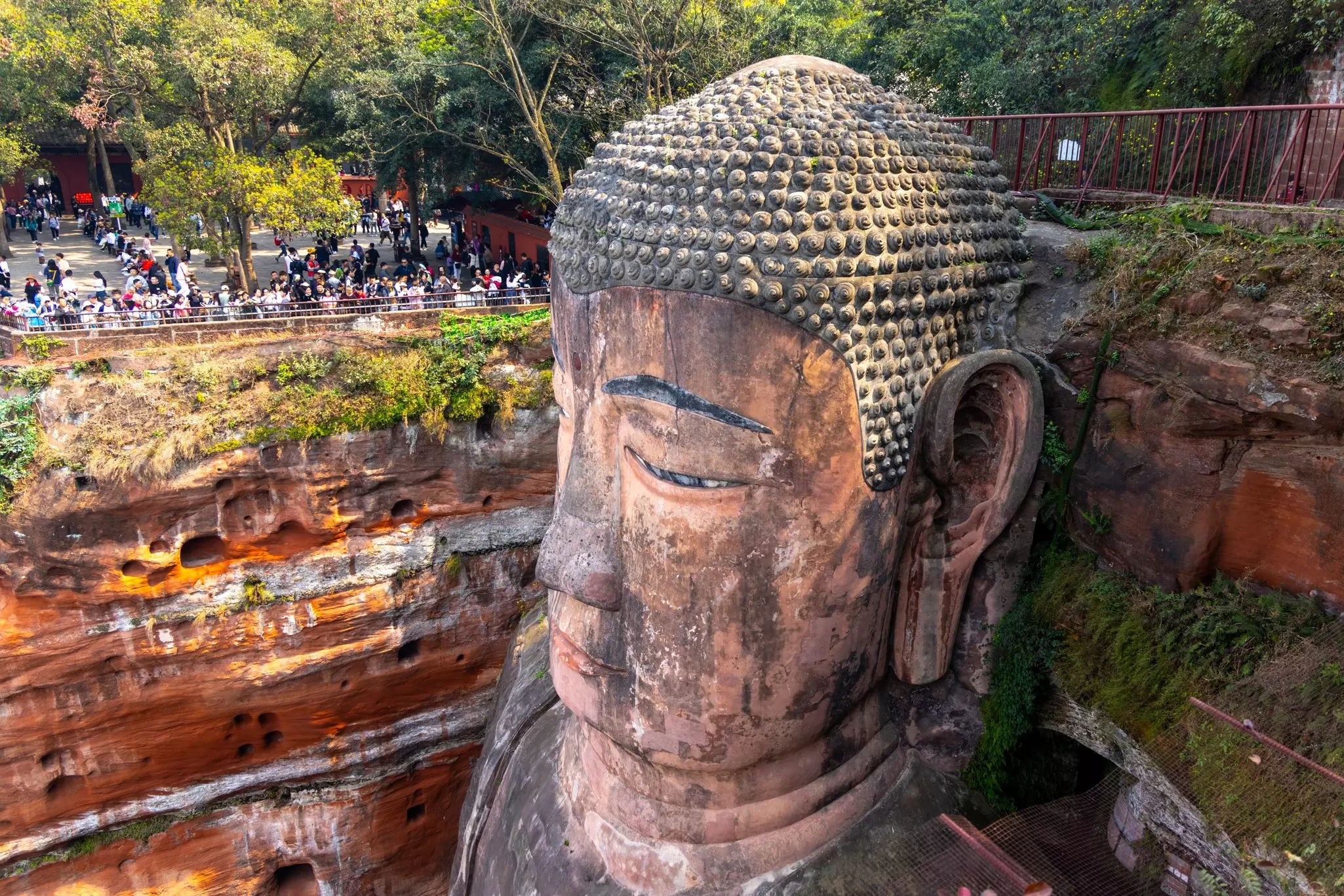 A monumental head of the Buddha is sculpted into a rock face on a cliff. Dozens of peopke, who appear tiny, gather on a platform to look on.