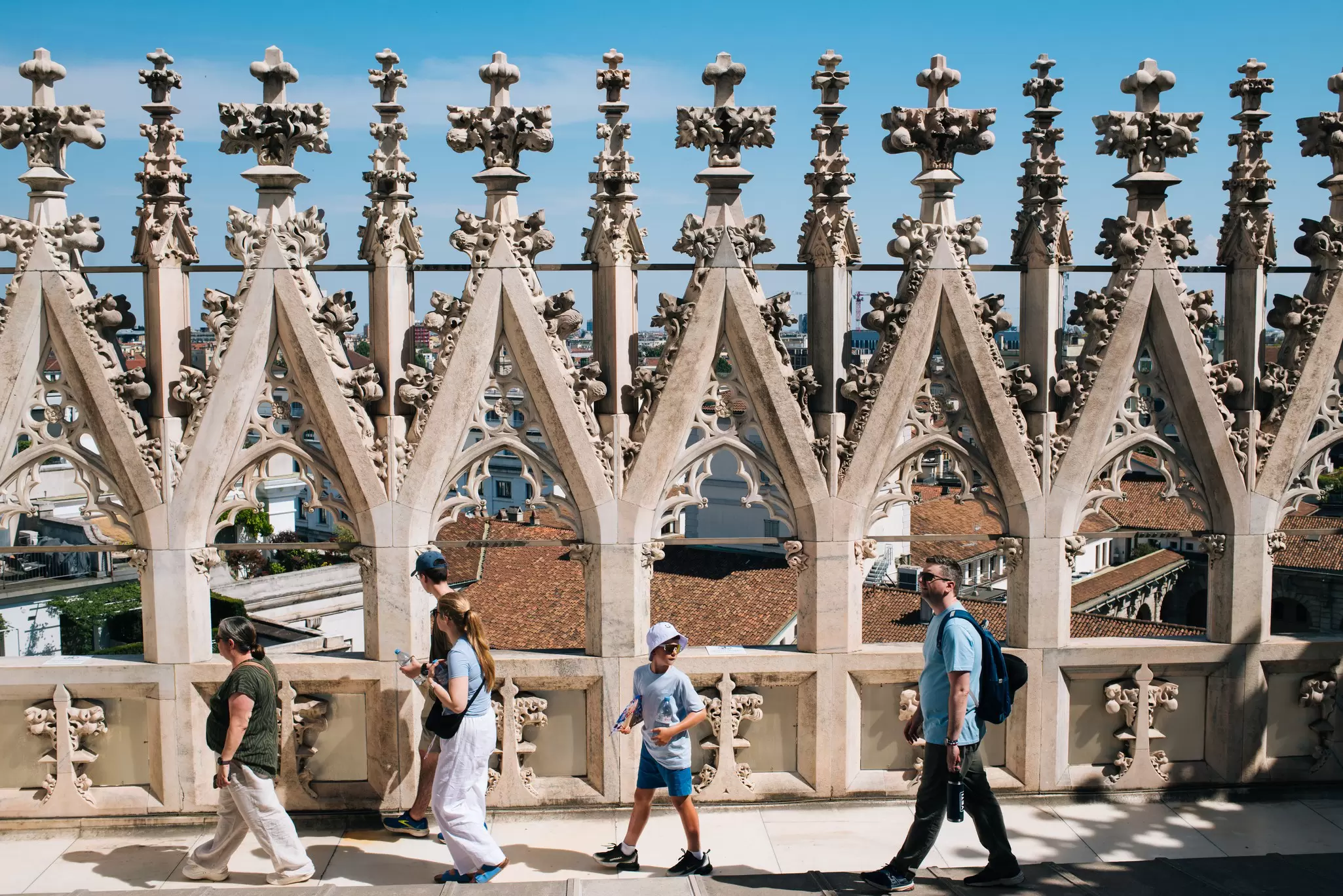 Visitors walking across the roof of a cathedral with intricate Italian Gothic pillars. A red-roofed city is visible from the rooftop.