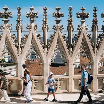 Visitors walking across the roof of a cathedral with intricate Italian Gothic pillars. A red-roofed city is visible from the rooftop.