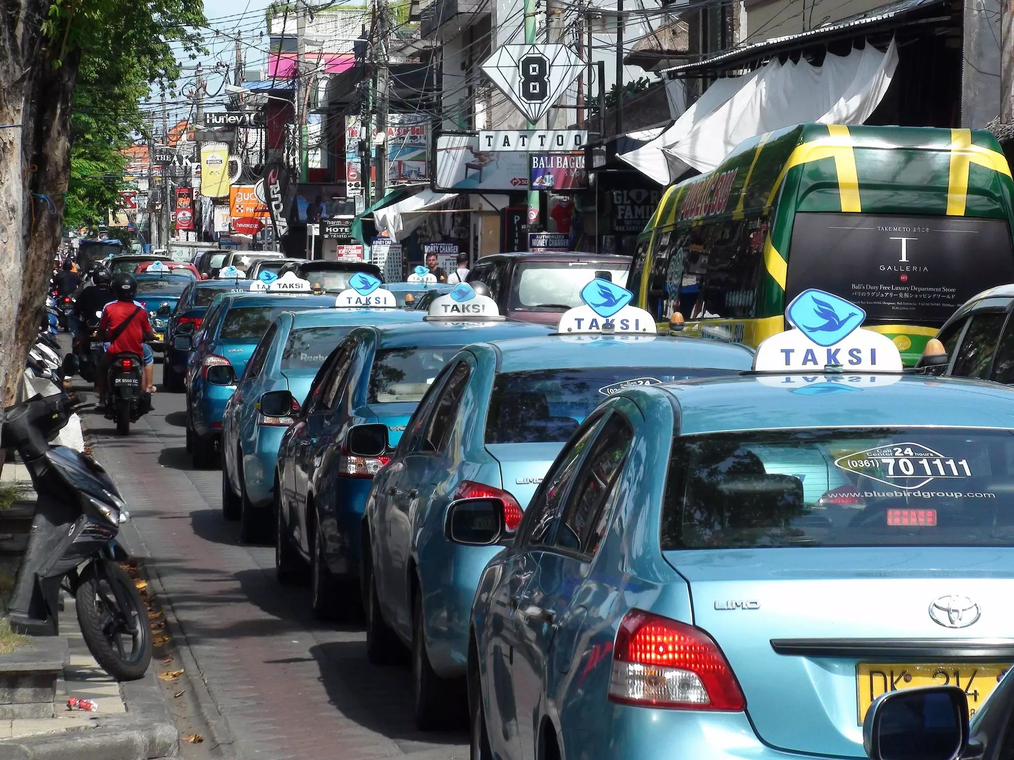 A busy street with a road full of waiting cars. Seven blue taxis stand waiting in a line.