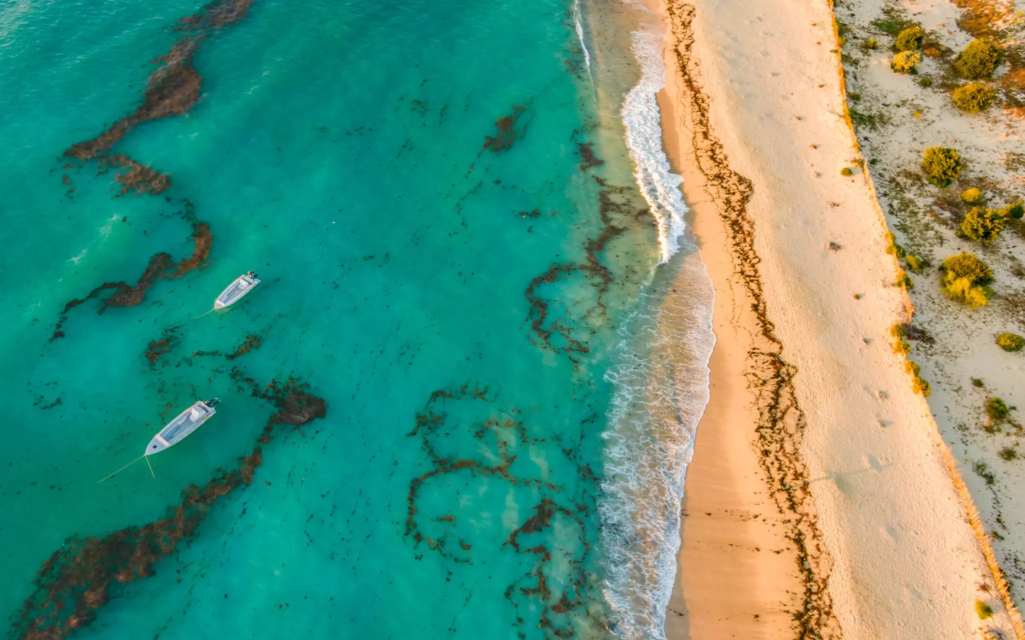 An aerial view of a tropical beach with waves from the turquoise-colored sea washing up on golden sand. Two boats are moored in the water just offshore.