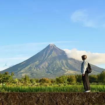 Southeast Luzon's Mayon Volcano rising over green rice fields, a traveller on a stone wall and HELLO MAYON letters at left