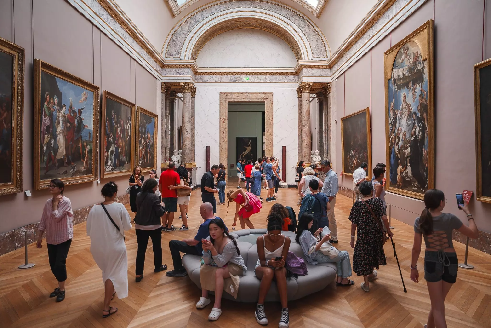 Brightly lit interior of a gallery at the Louvre Museum in Paris, France, filled with a large group of people.