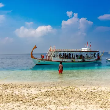 A man stands in the shallows of the ocean near the edge of a sandy beach to direct a tour boat in Fulidhoo, Maldives