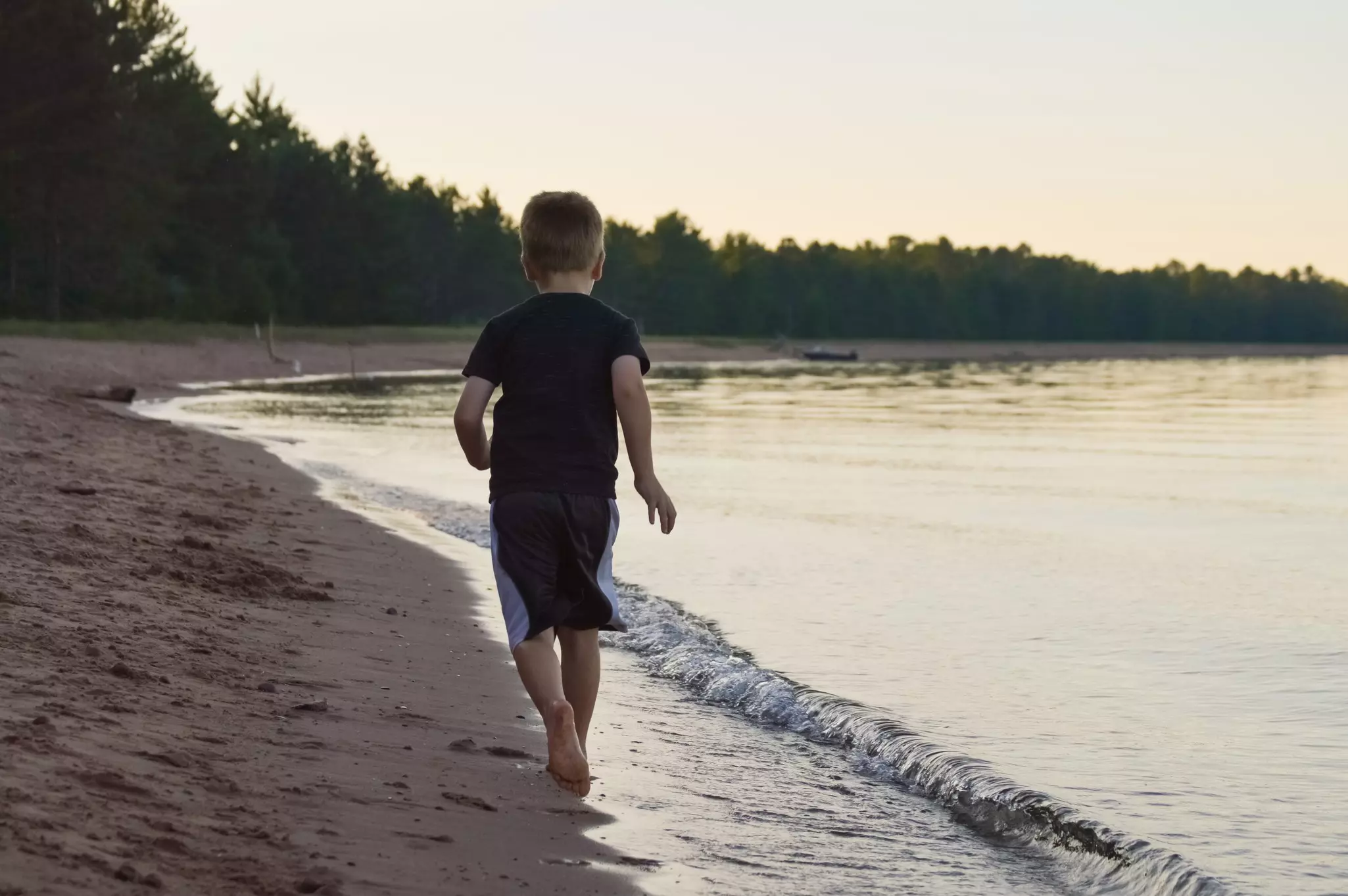 School aged boy runs on a deserted sandy beach surrounded by evergreen forest with a boat beached in the distance under yellow evening sky, Northern Wisconsin, Great Lakes, USA