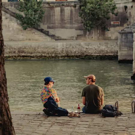 Two people sat on a riverbank at sunset with a bottle of wine between them
