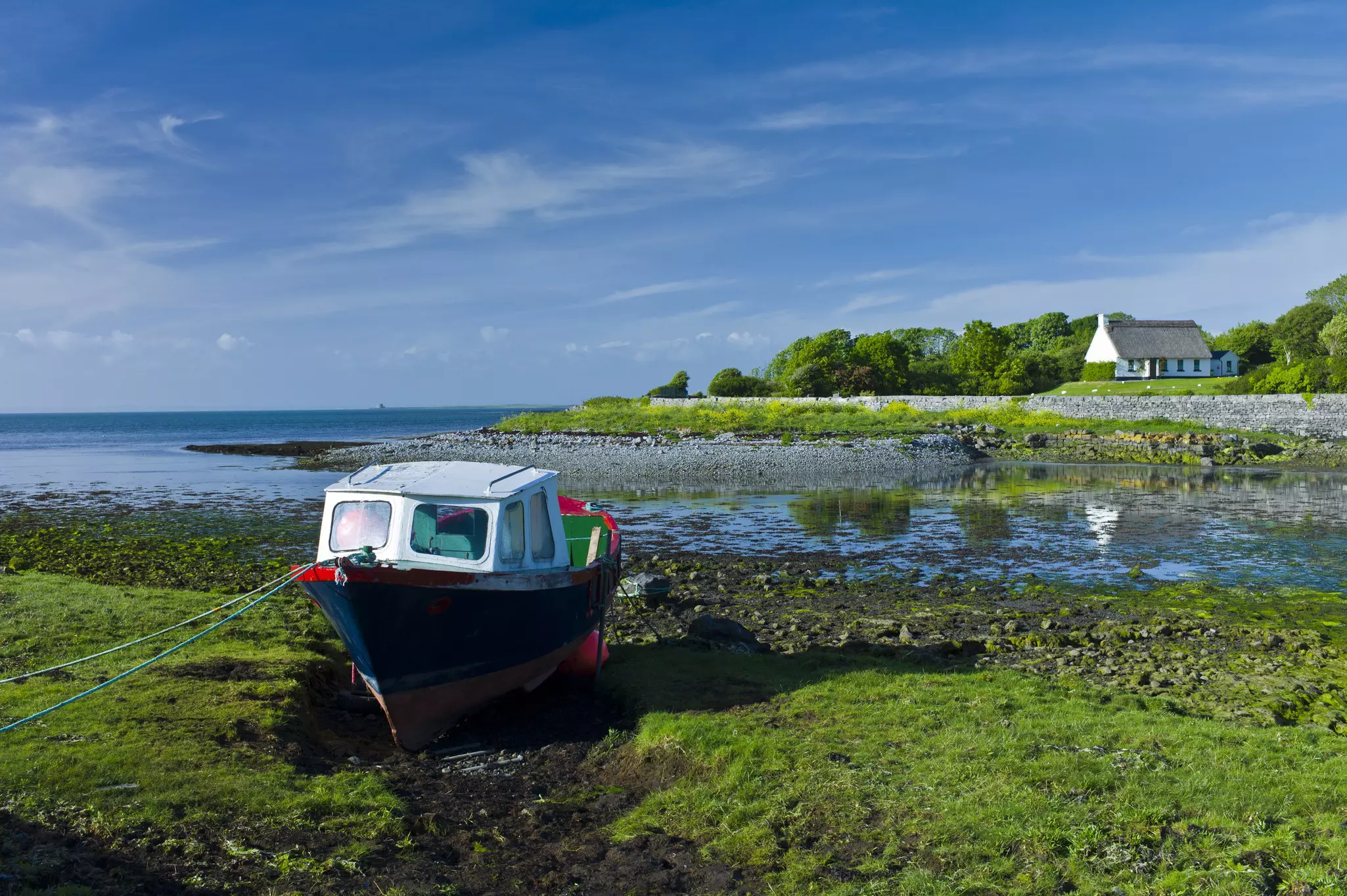 A fishing boat is docked on a grassy bank. A thatched cottage sits on the other side of the water.