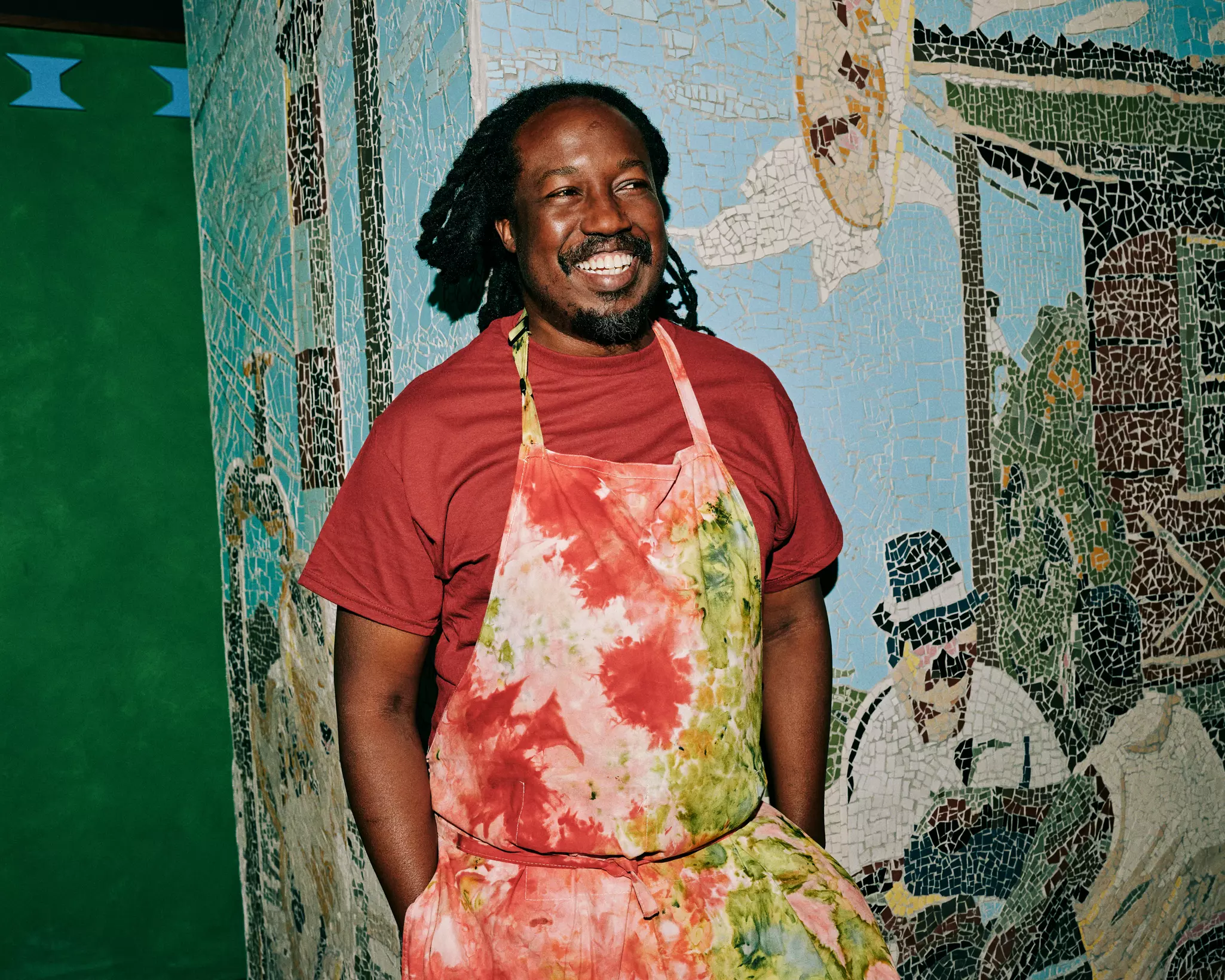 A smiling man wearing a red T-shirt and a tie-dye apron stands in front of a mosaic mural.