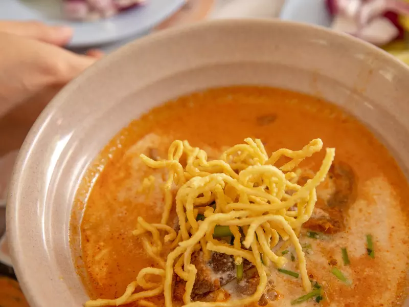 A bowl of rich red soup in a bowl, with fried noodles on top and toppings in the background. 