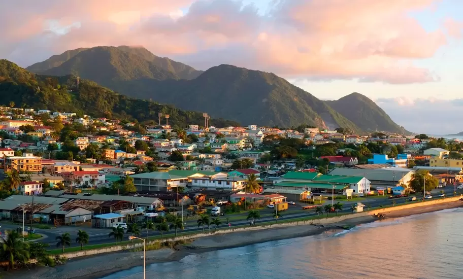 Hillside houses of Roseau, Dominica, along a seafront road below forested mountains under pink evening clouds