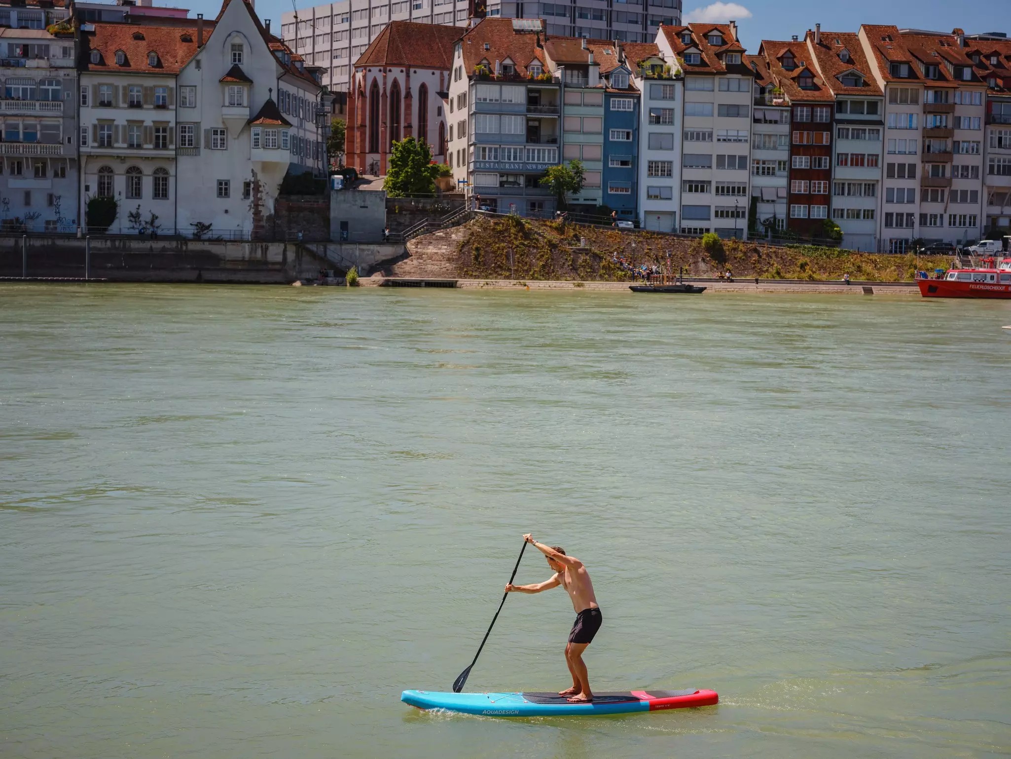 A man on a stand-up board floats on the river Rhine, in the city center of Bern, with apartment buildings along the shoreline