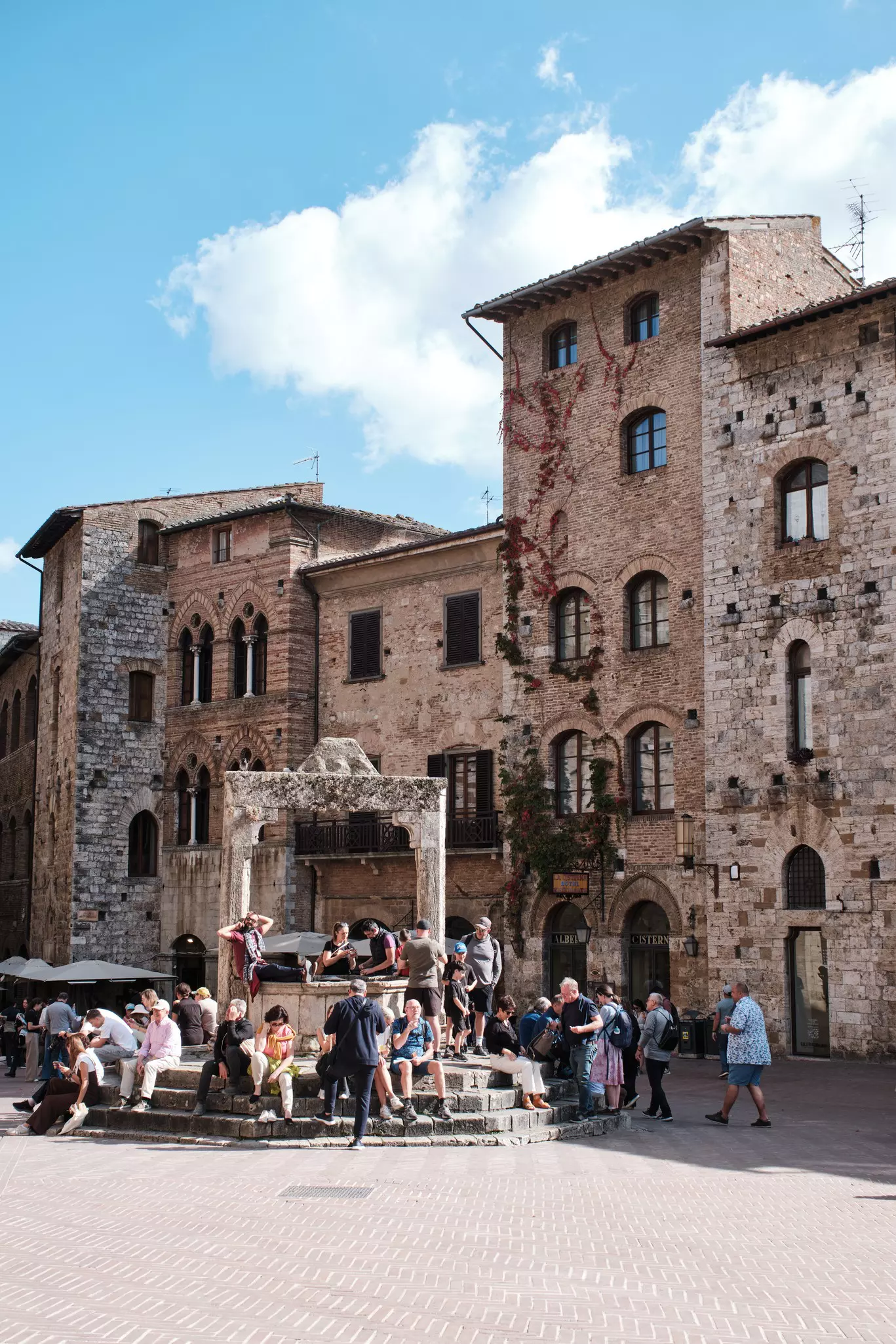 A square in a medieval city. People pause on the steps around an ancient stone cistern.