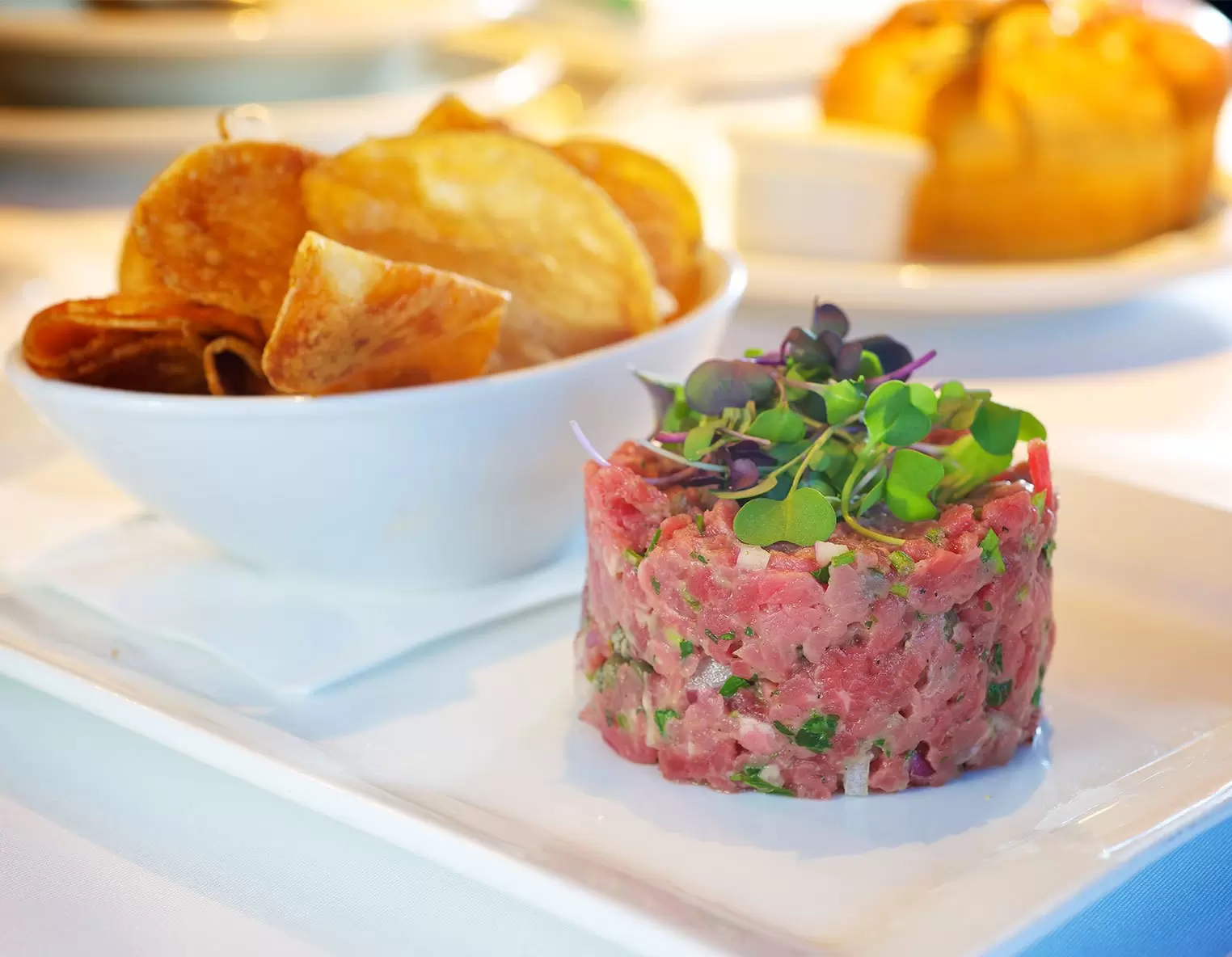 steak tartare topped with micro greens and a small bowl of potato chips on the side