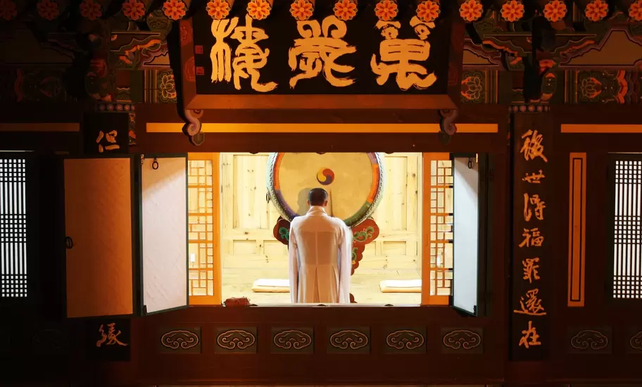 Monk in a white robe facing a large ceremonial drum through an open temple window, Korean characters on the beams above