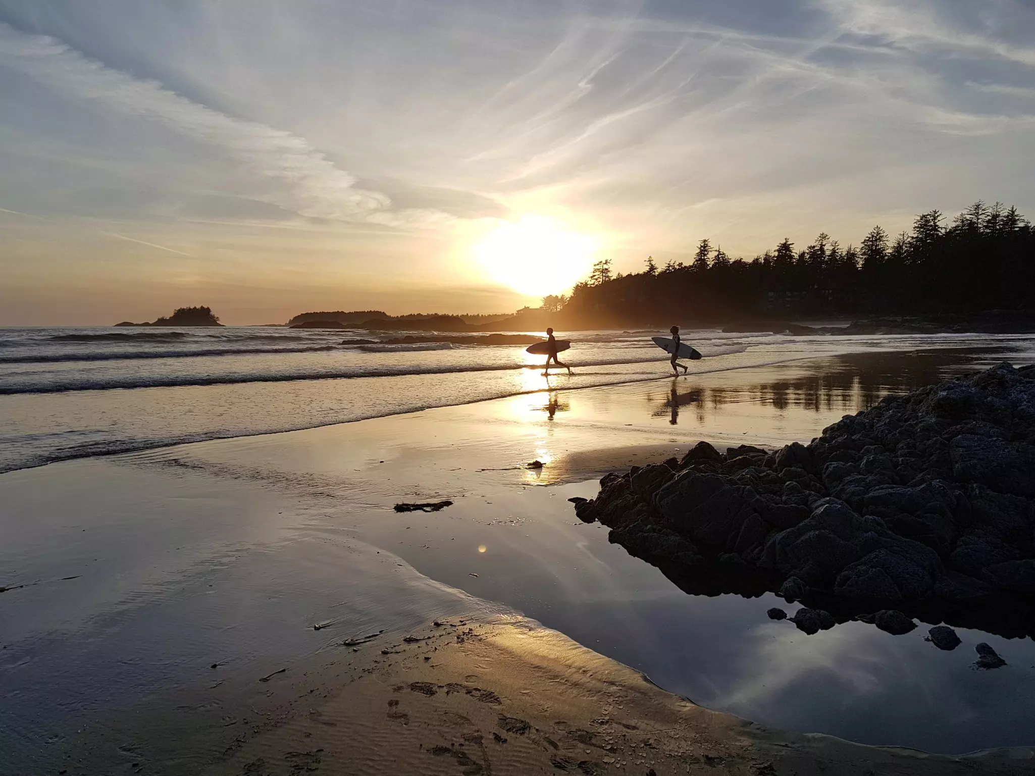 Two surfers carrying their boards and walking along a beach at low tide are silhouetted in the sunset light.