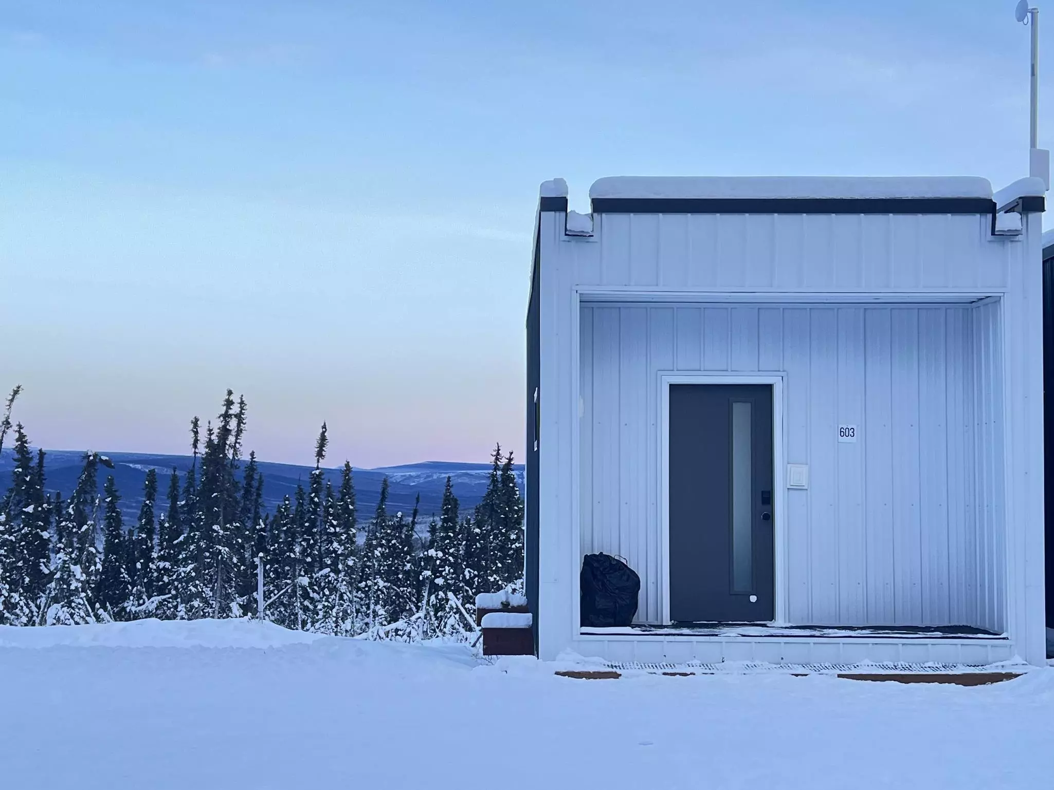 A square white building with a dark door in a snowy landscape with evergreen trees in Alaska.