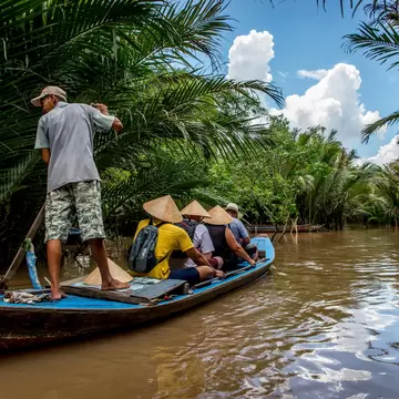 Man standing on the stern of a blue wooden boat carrying passengers in conical hats along a brown, palm-lined canal.