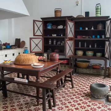 Wooden table and benches with a PLEASE DO NOT SIT sign, dark cabinets of pots and bowls, red star-patterned floor tiles